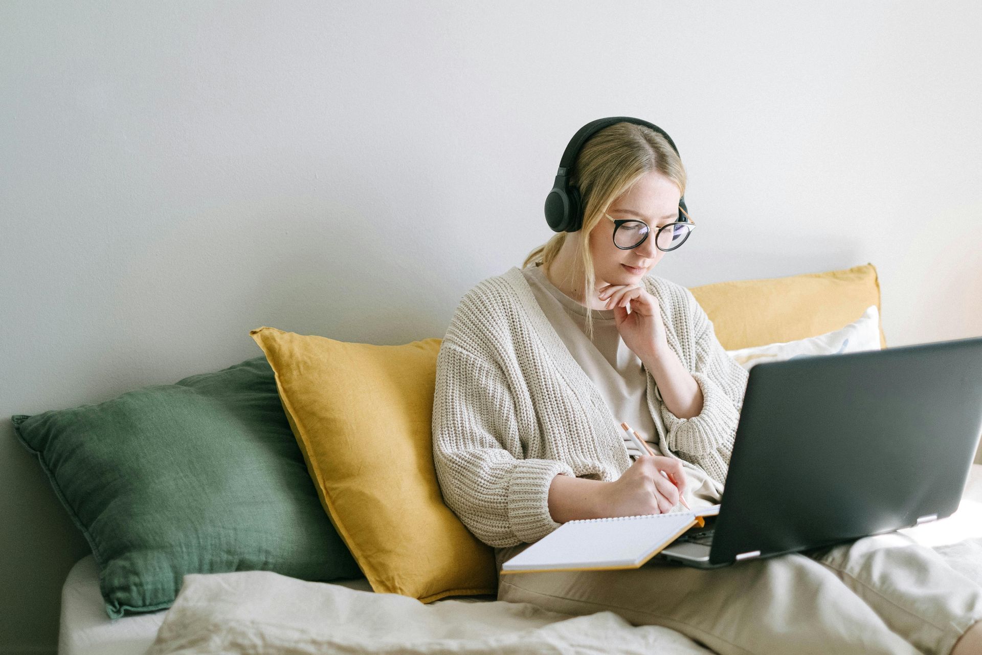 Person working on a laptop on a couch, wearing headphones and writing in a notebook.