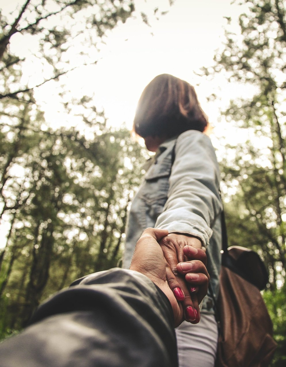 Person holding hands with a companion while walking in a wooded area, viewed from behind.