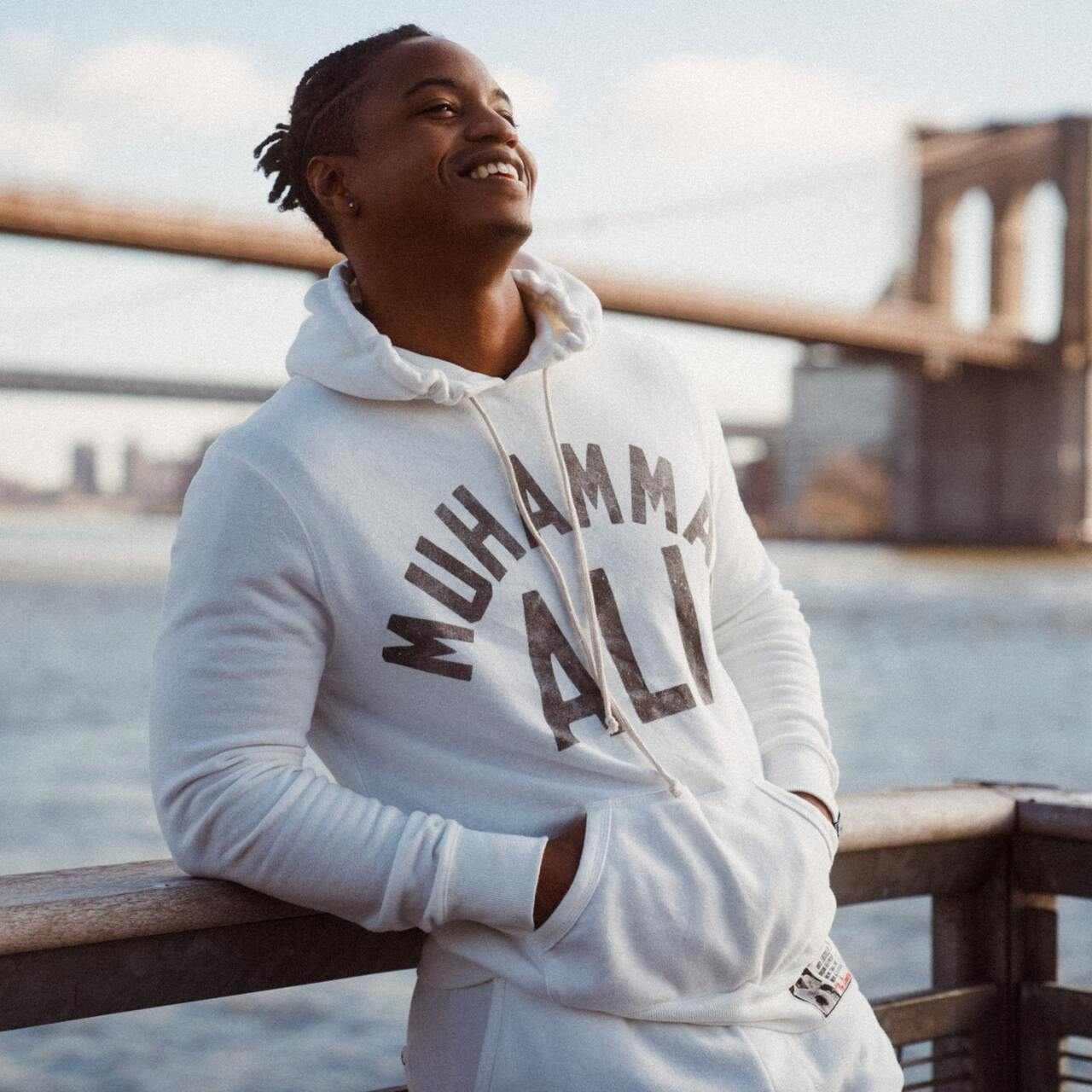 Person in a white hoodie leaning on a railing by the water with a bridge in the background.