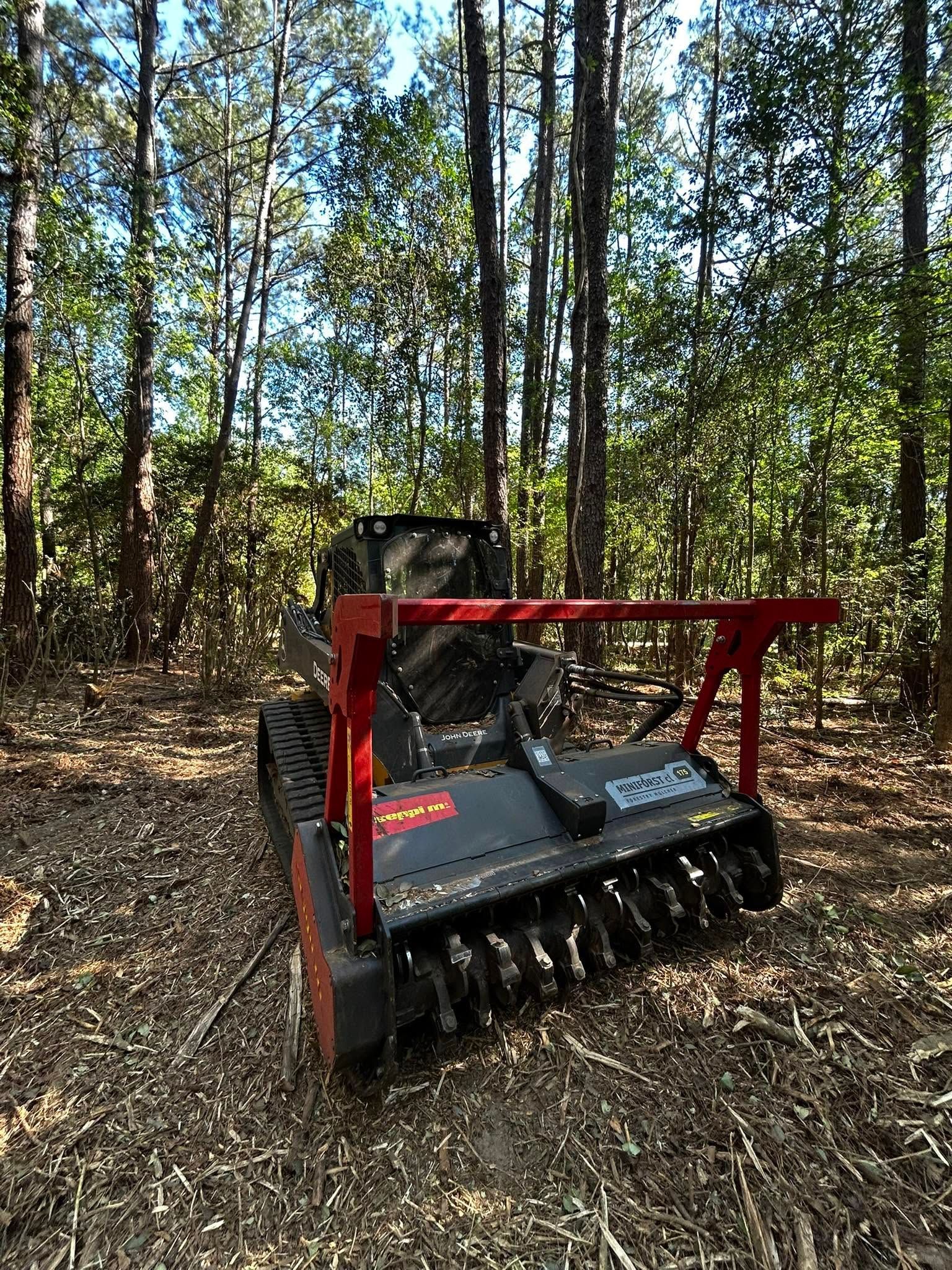 Red forestry mulcher attachment in a wooded area with trees and leaf-covered ground