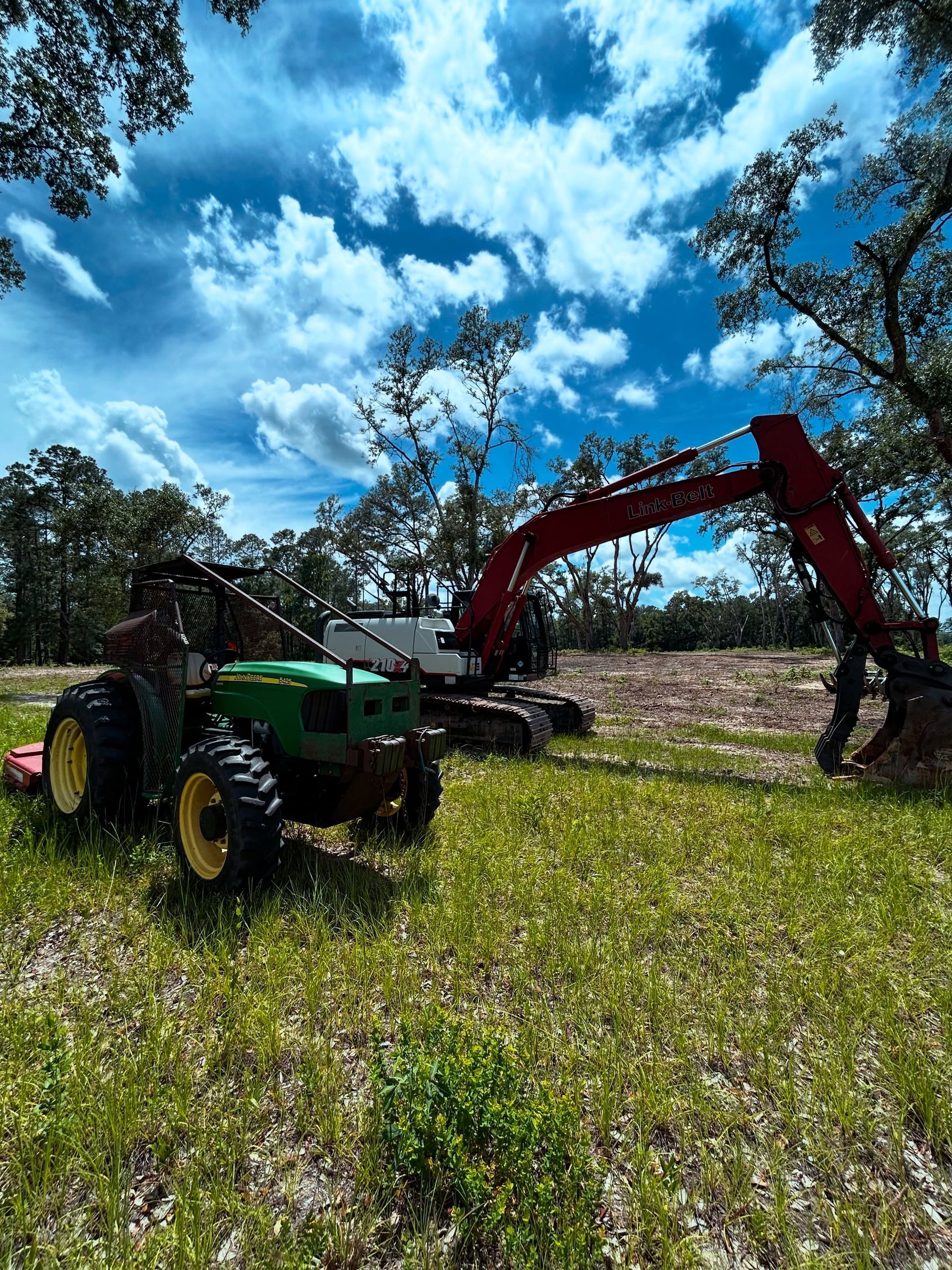 Green tractor and red excavator working in a grassy field under a bright blue sky.