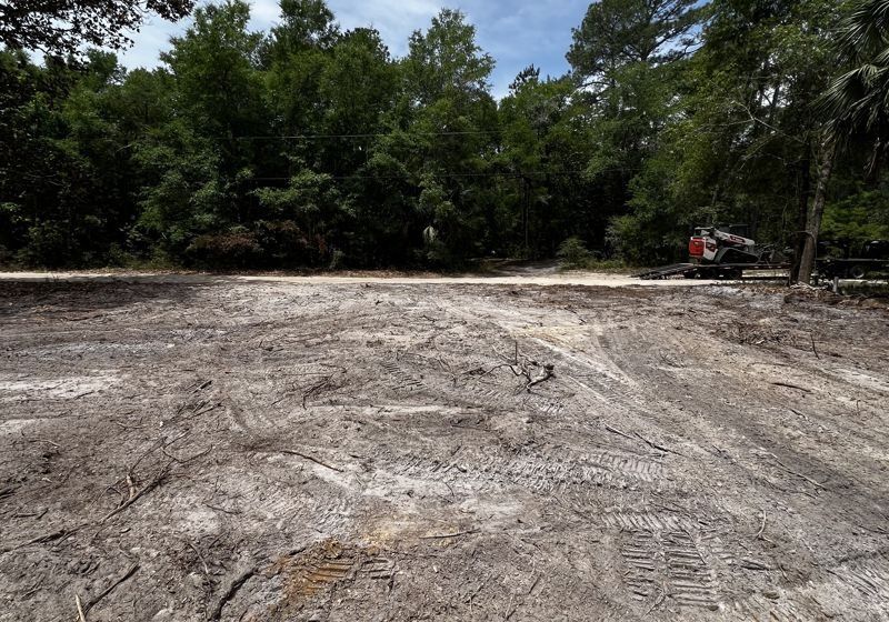 Cleared dirt lot bordered by trees, with a small excavator working on the right side.