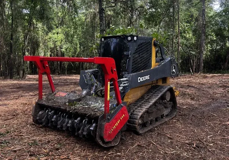 Black skid-steer mulcher with red attachment clearing brush in a wooded forest area