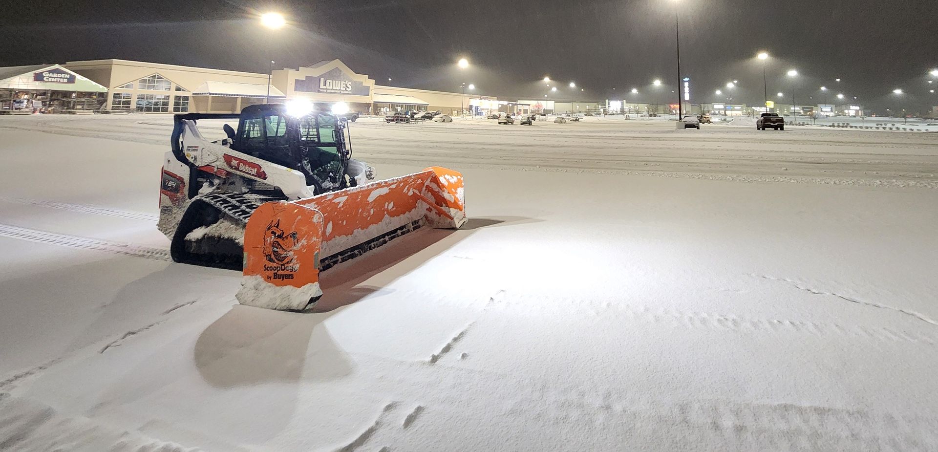 Orange snowplow clearing a snow-covered road at night under bright streetlights