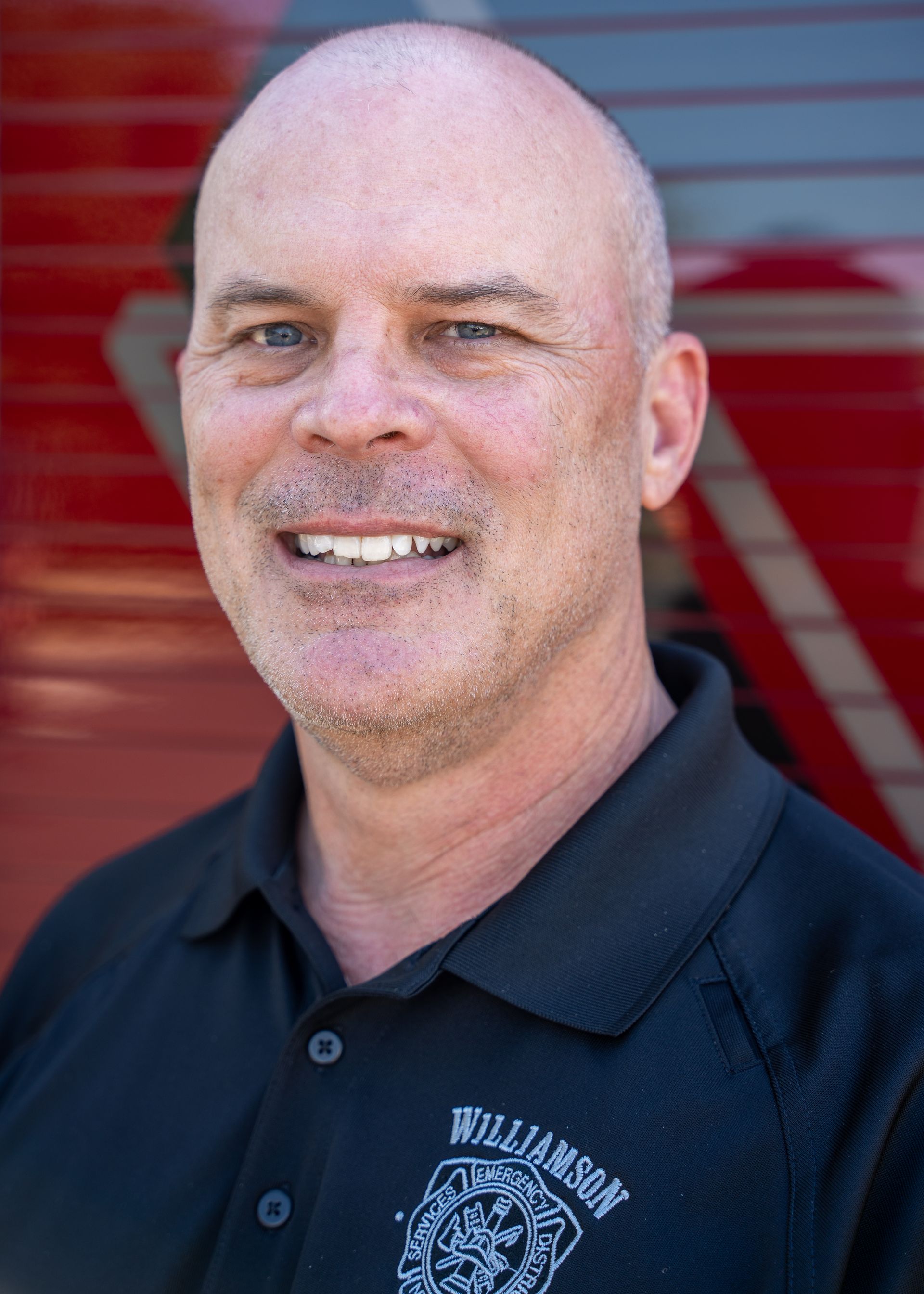 A bald man wearing a black shirt is smiling in front of a red truck.