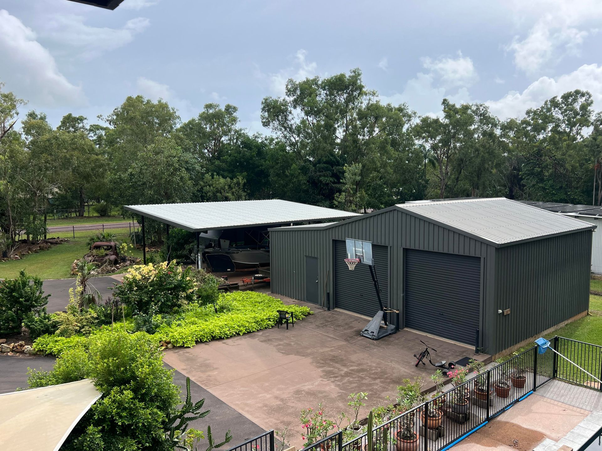 Two sheds with silver roofs and dark green walls on a concrete pad with a basketball hoop. Trees in background — AKW Builders Pty Ltd In Leanyer, NT