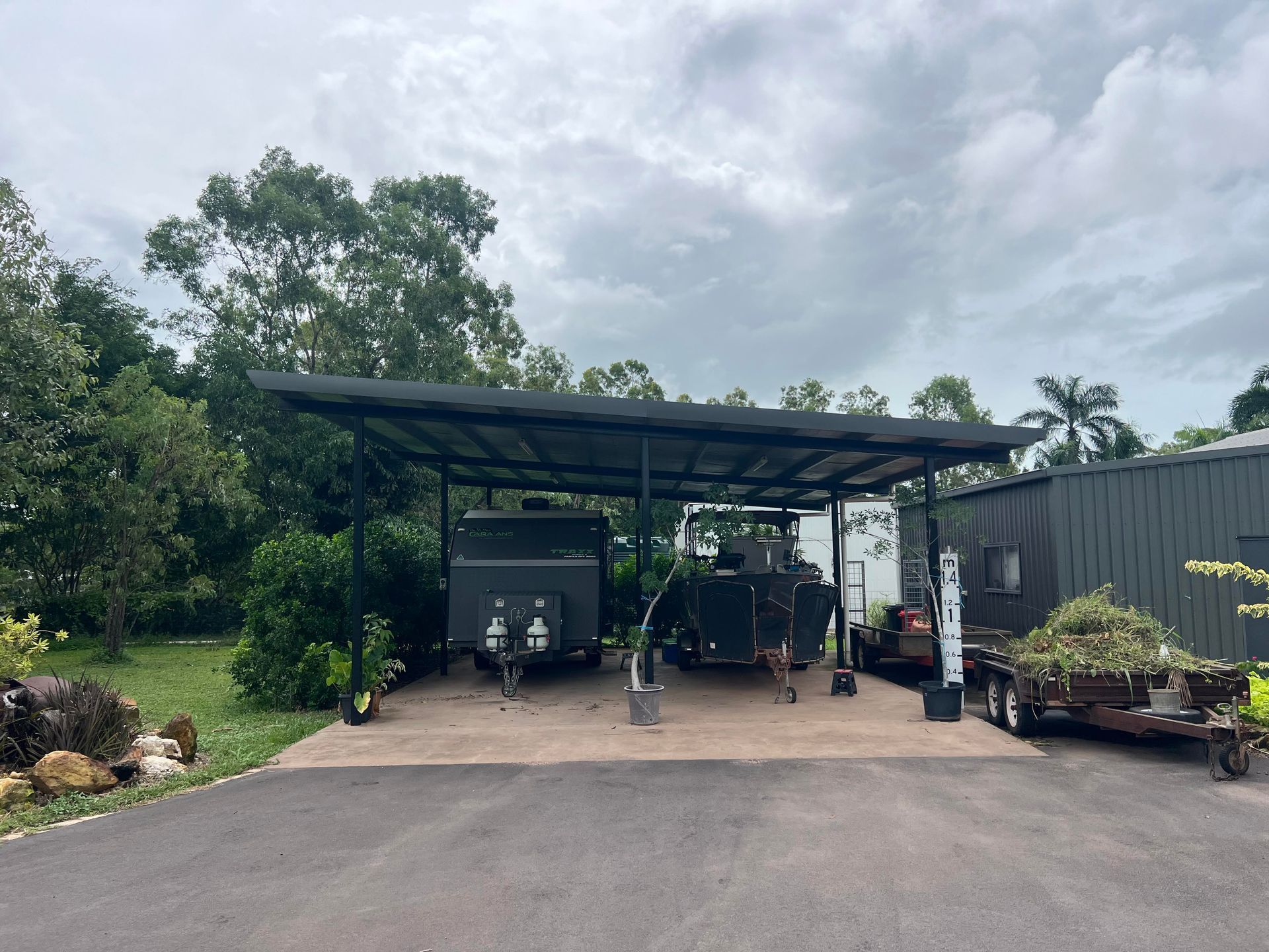 A covered carport with a trailer, boat and trailer on a concrete slab. Green foliage and a building are nearby — AKW Builders Pty Ltd In Leanyer, NT