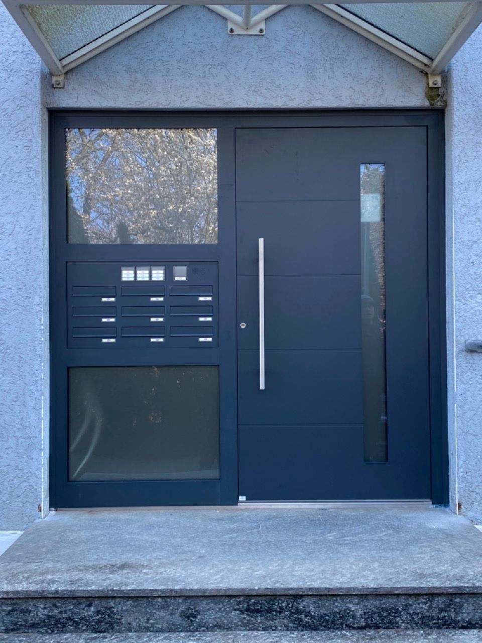 Moderne dunkelblaue Haustür mit Briefschlitzen und vertikalem Türgriff in einem grauen Stuckeingangsbereich.