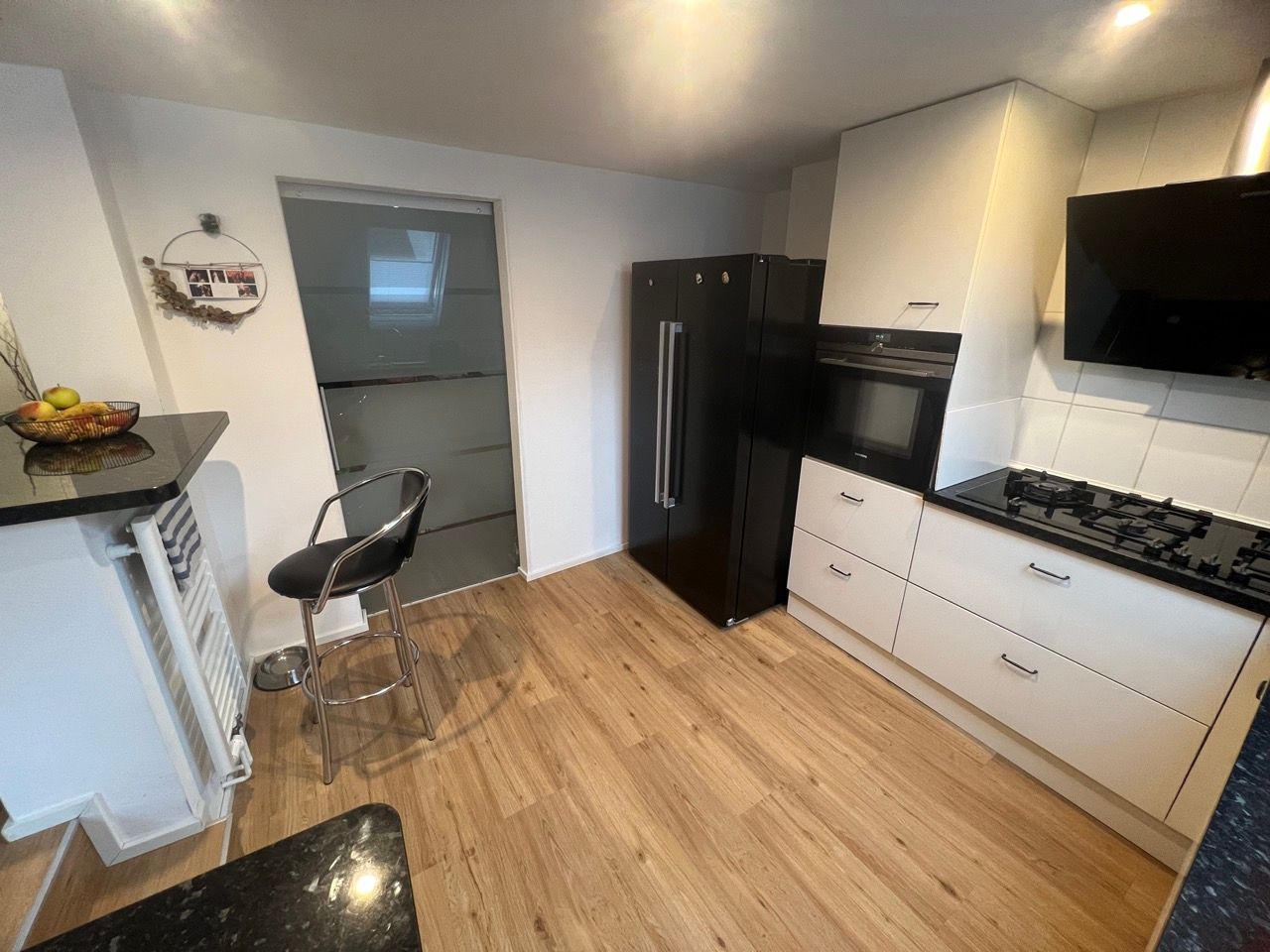 Moderne Küche mit Holzboden, weißen Schränken, schwarzen Geräten und einem kleinen Hocker an der Küchentheke.