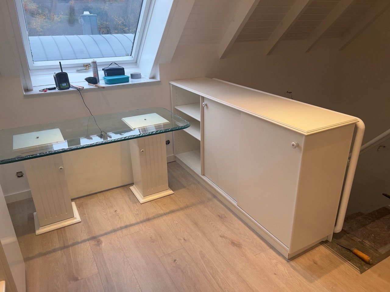 Kleines Zimmer mit einem Glastisch am Fenster und einem weißen Klapptisch an der Wand.