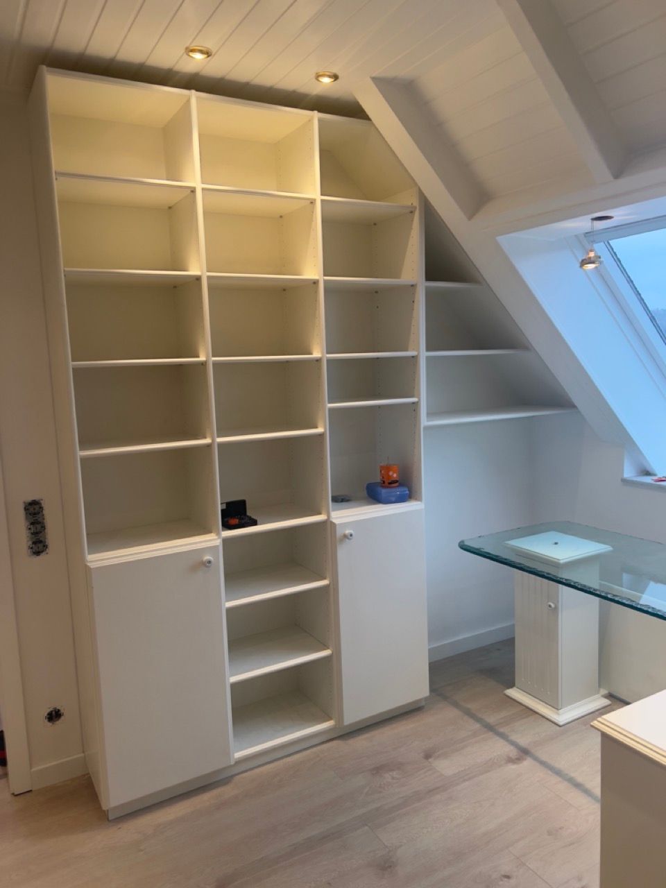 Eingebaute weiße Regale in einem hellen Dachzimmer mit Oberlicht und Glasschreibtisch