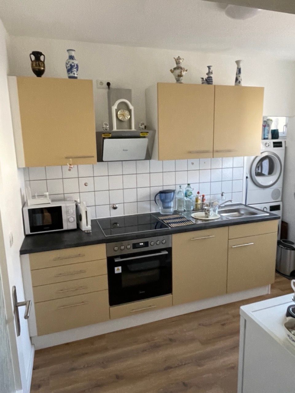 Kleine Küche mit beigen Schränken, schwarzer Arbeitsplatte, weißem Fliesenspiegel und Holzboden