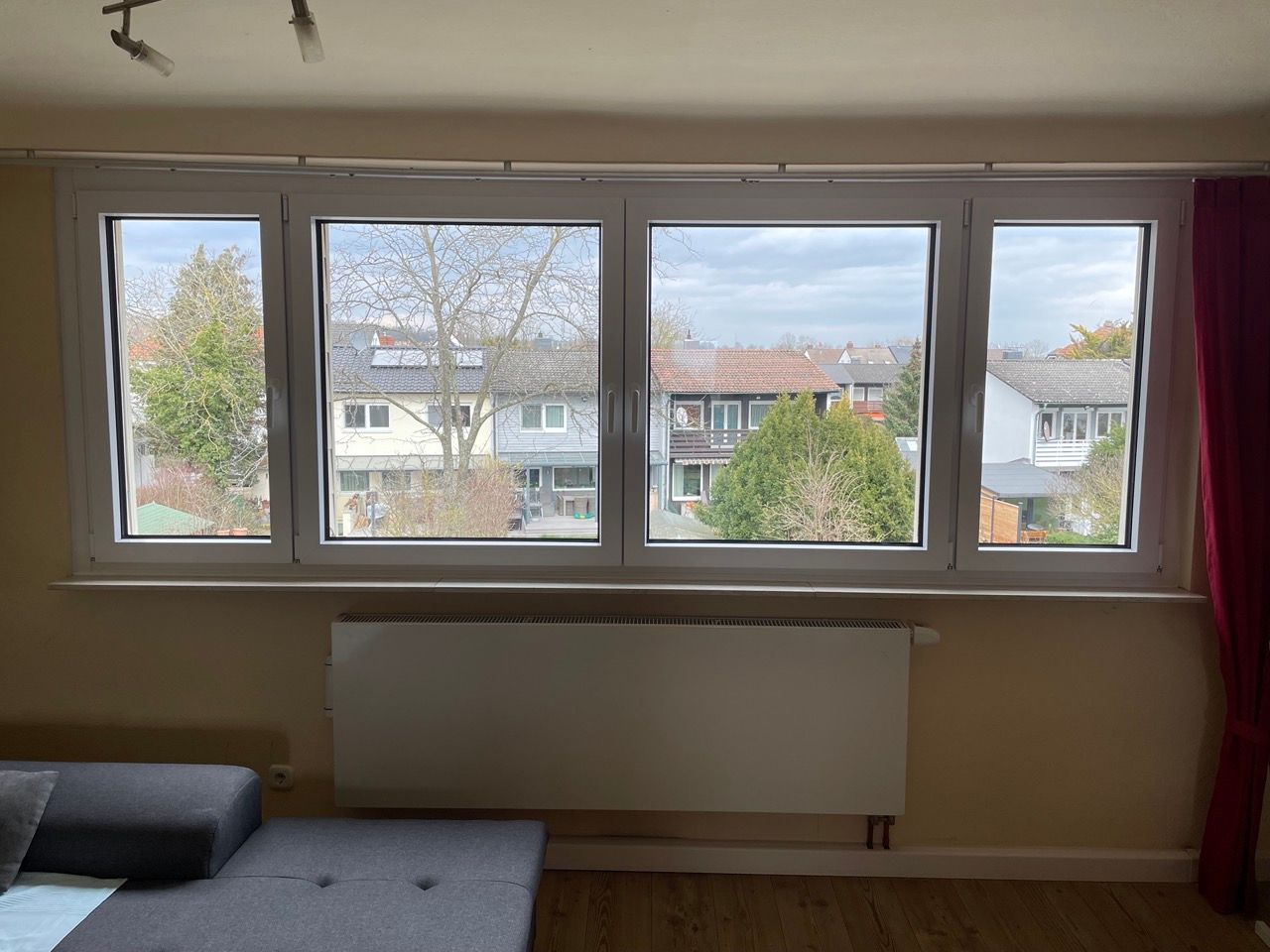 Wohnzimmer mit großem Fenster, darunter ein Heizkörper und Blick auf Häuser und Bäume draußen