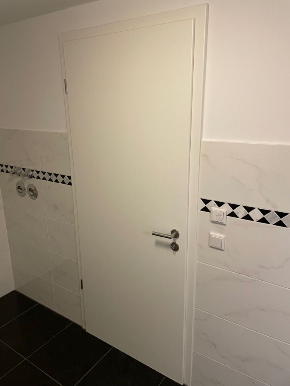 Weiße Tür in einem gefliesten Badezimmer mit schwarzen Bodenfliesen und einer dekorativen schwarz-weißen Bordüre