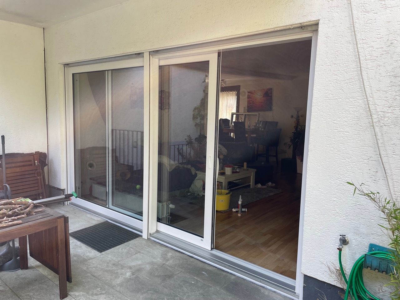 Schiebetüren aus Glas öffnen sich zu einer Terrasse und einem Wohnzimmer, in dessen Nähe ein Tisch und Topfpflanzen stehen.
