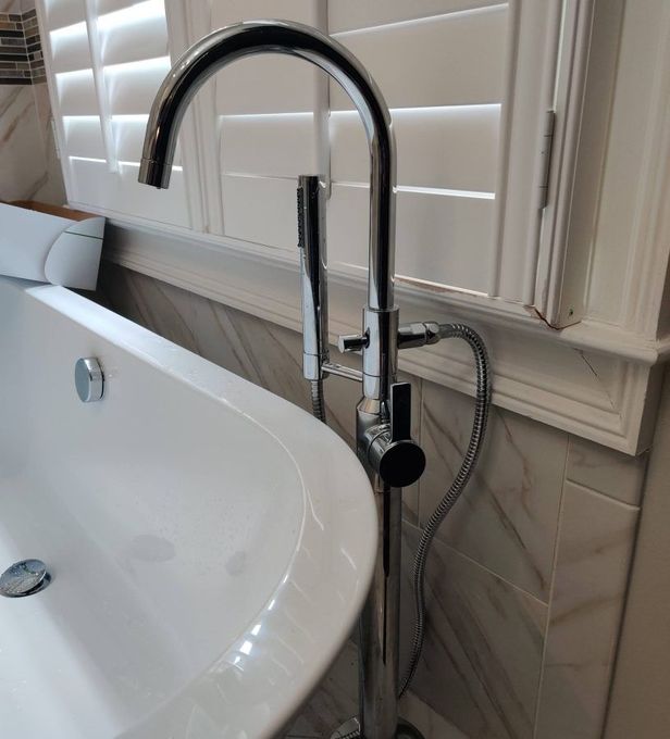 Chrome freestanding bathtub faucet next to a white tub and shuttered window.