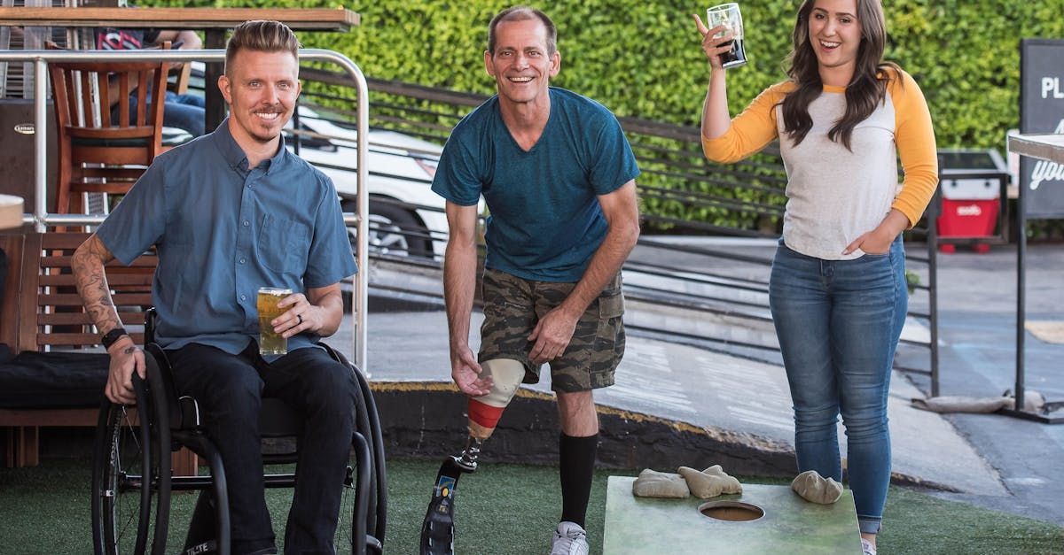 Three people outside, clinking glasses. Man in wheelchair and man with prosthetic legs smile. Woman cheers, holding drink.