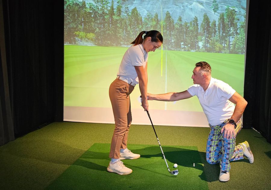 Two people practicing golf indoors, one kneeling to help the other hold a club on a green turf mat.