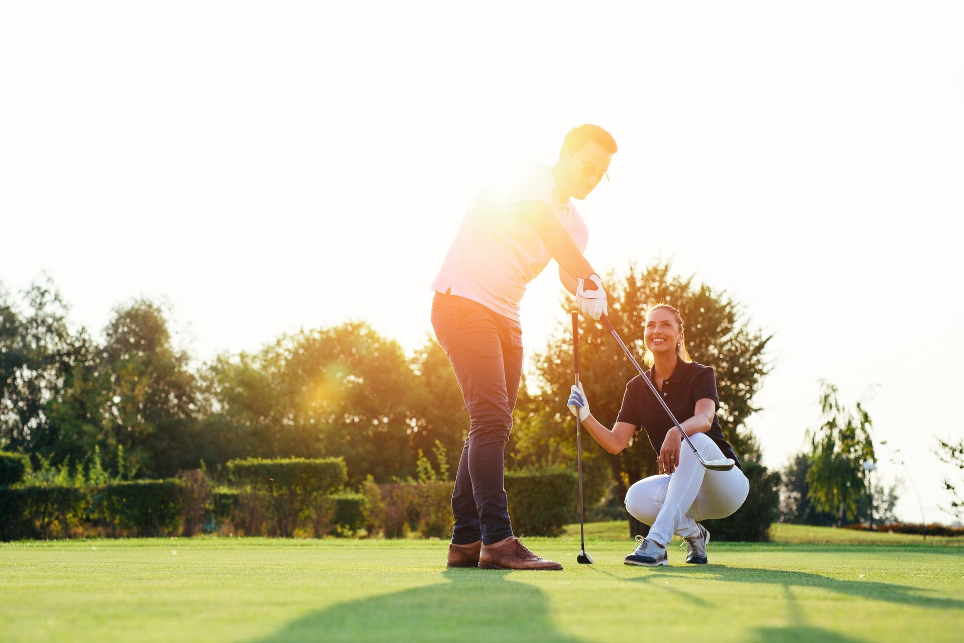 Two golfers on a green, one preparing to putt as the other kneels nearby in bright sunlight