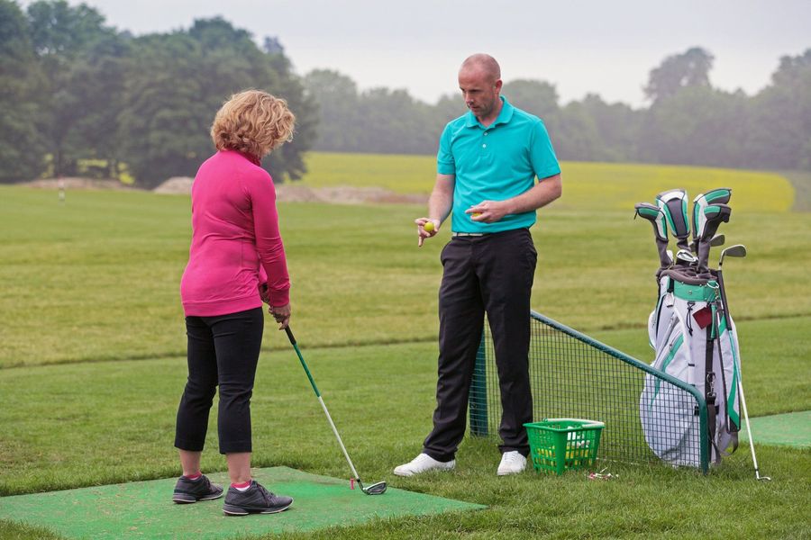 Golf instructor demonstrating a shot to a golfer on a practice range, with clubs and bucket of balls nearby.