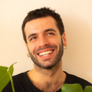 Smiling man with dark hair and earrings, holds plant, against a light background.