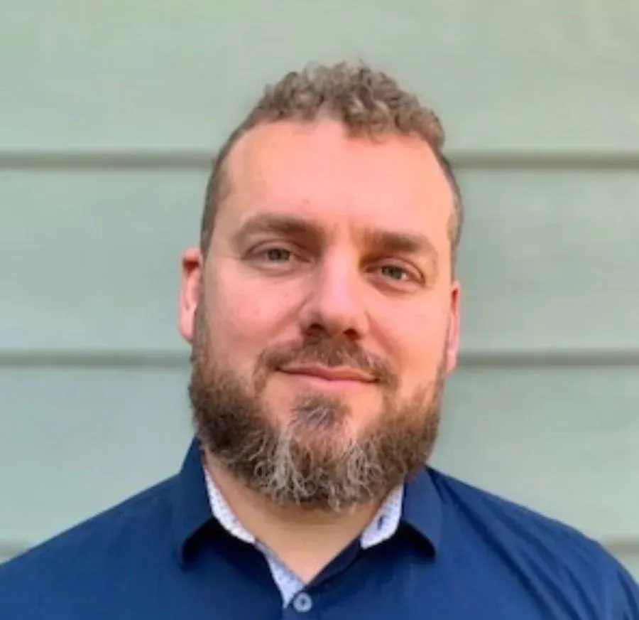 Man with a beard and curly hair smiles in front of a teal wall, wearing a blue shirt.