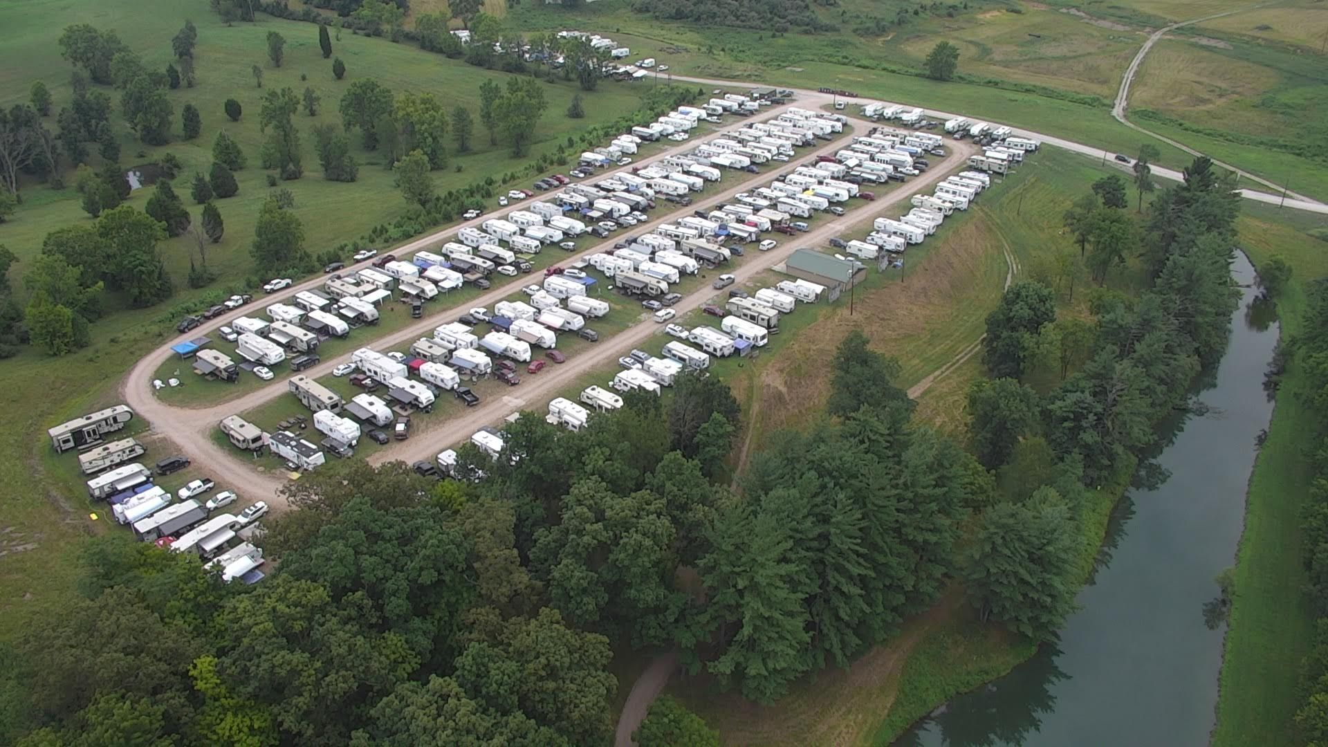 An aerial view of an RV park with rows of recreational vehicles parked in a grassy field next to a river and forest.