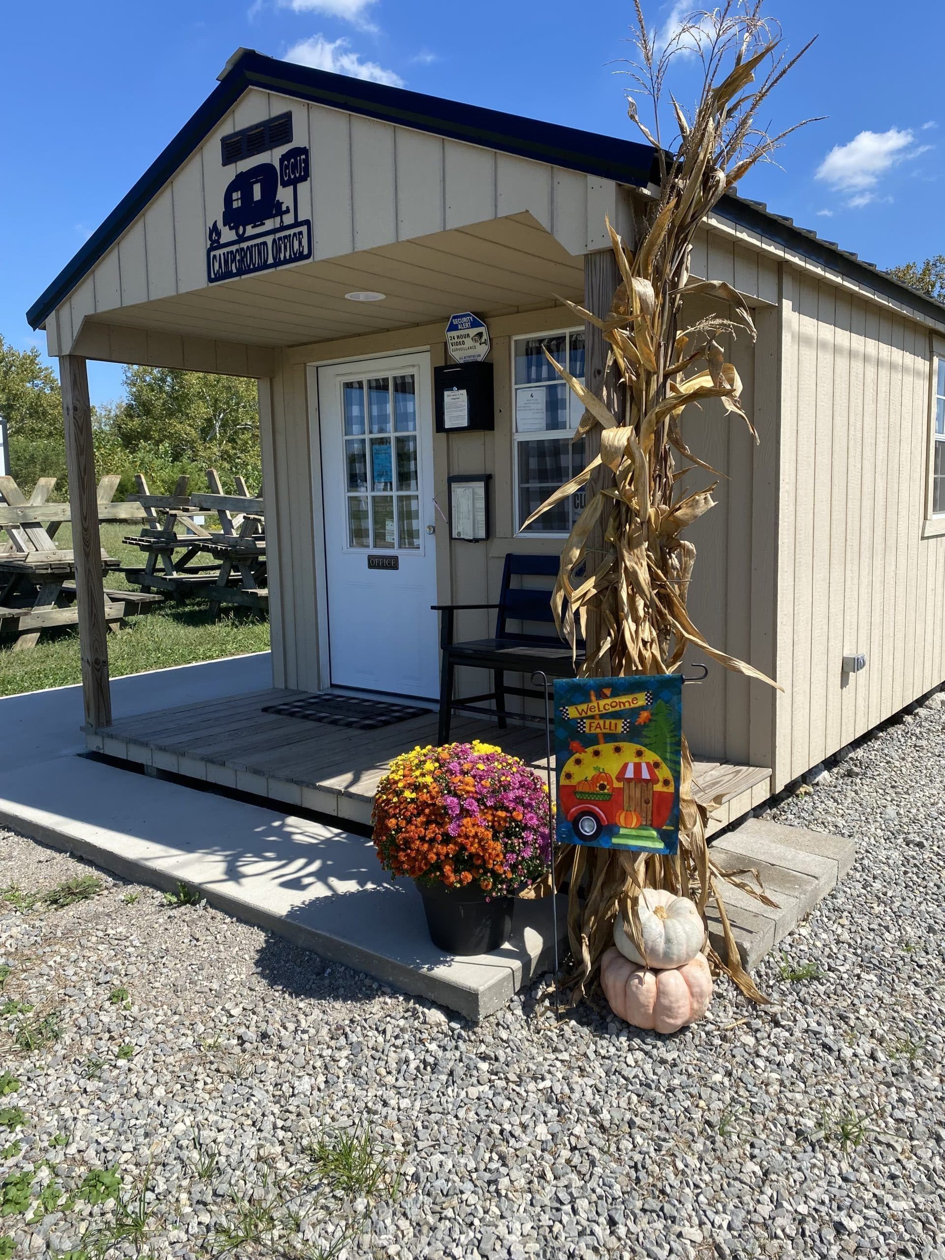 A beige wooden shed decorated for autumn with a corn stalk, potted mums, and small pumpkins on the front porch.