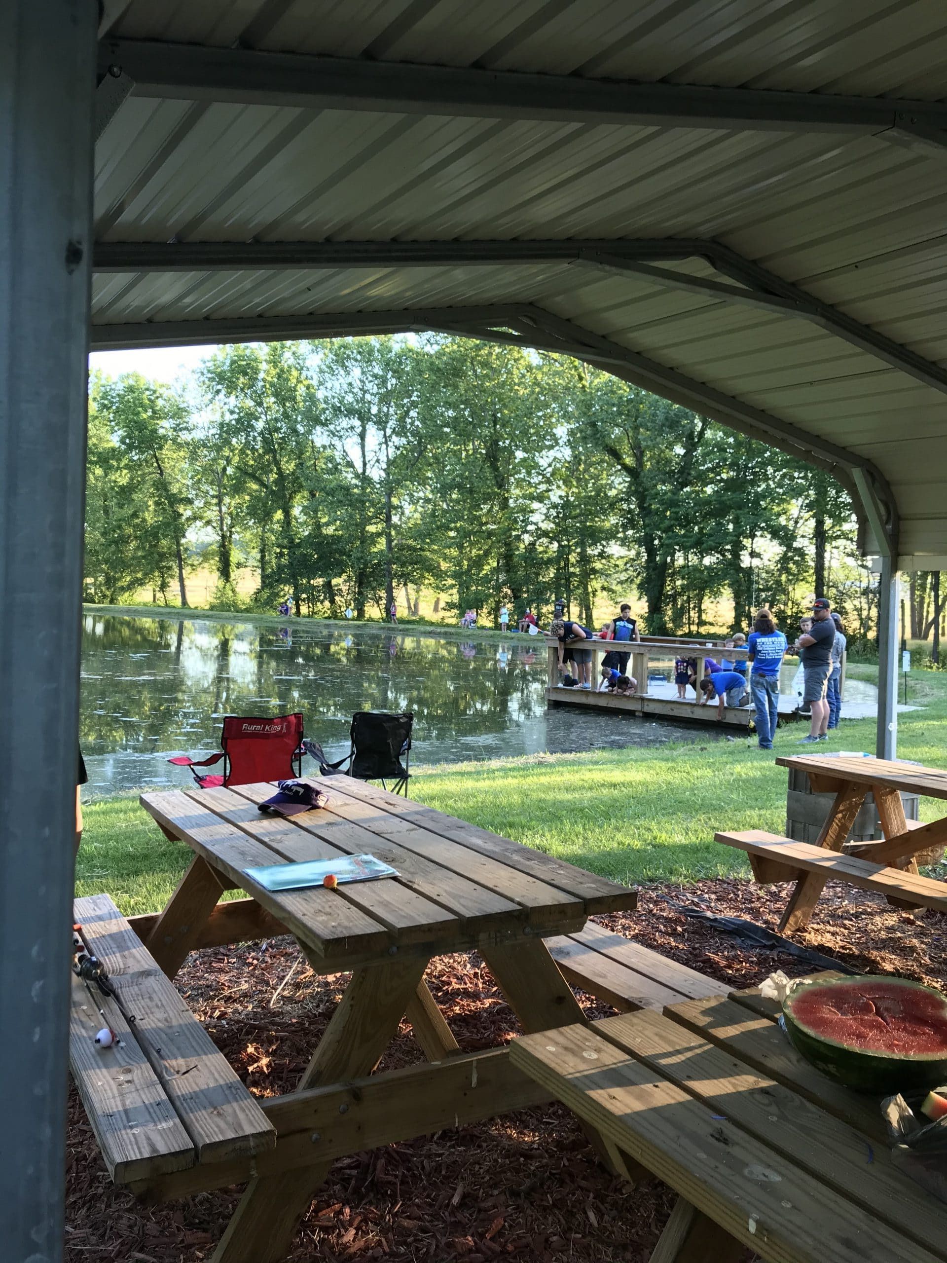 Picnic tables under a pavilion overlook a pond with people standing on a wooden dock among trees in the background.