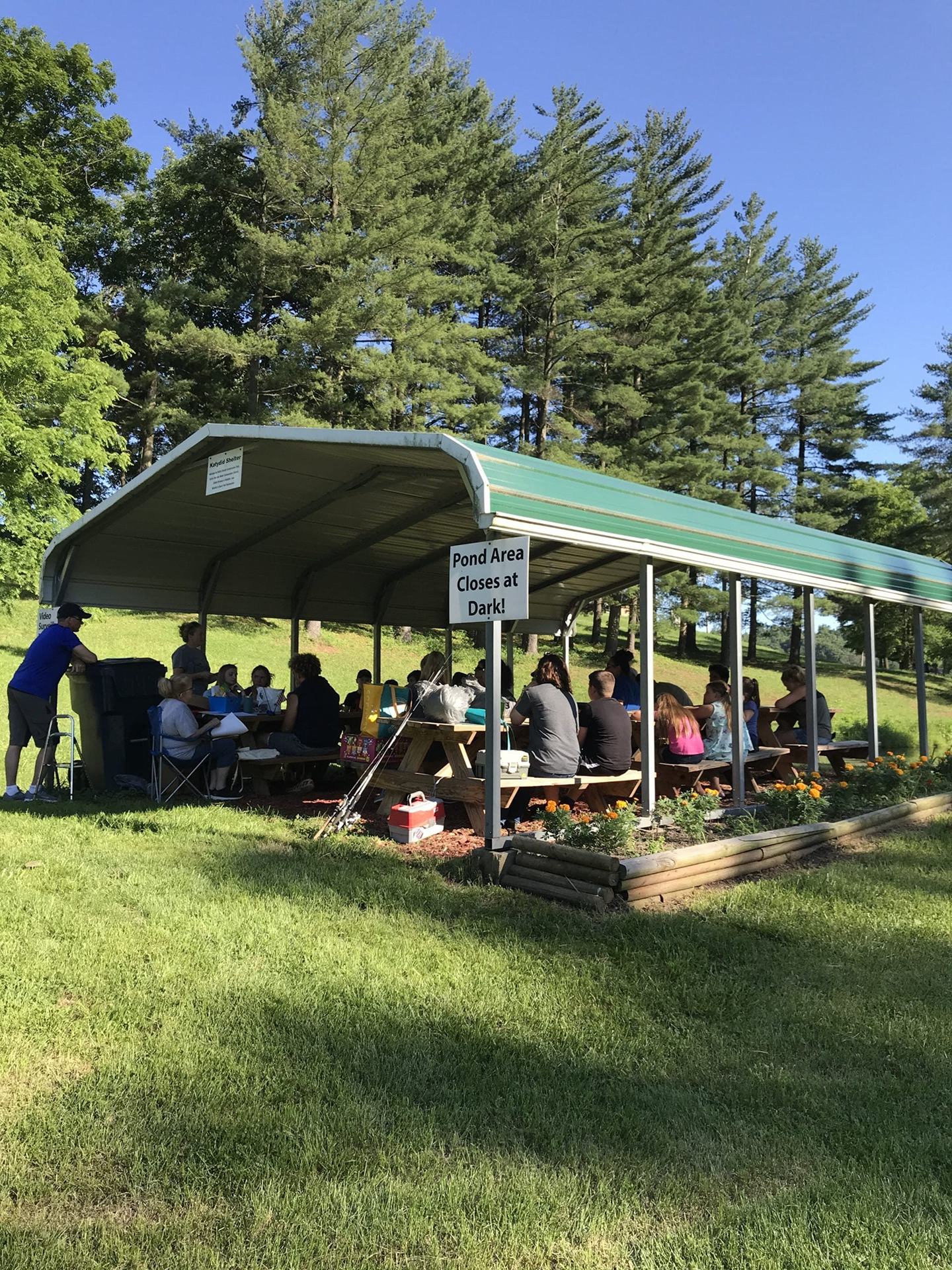People gather at picnic tables under a green canopy structure in a grassy park setting with trees in the background.