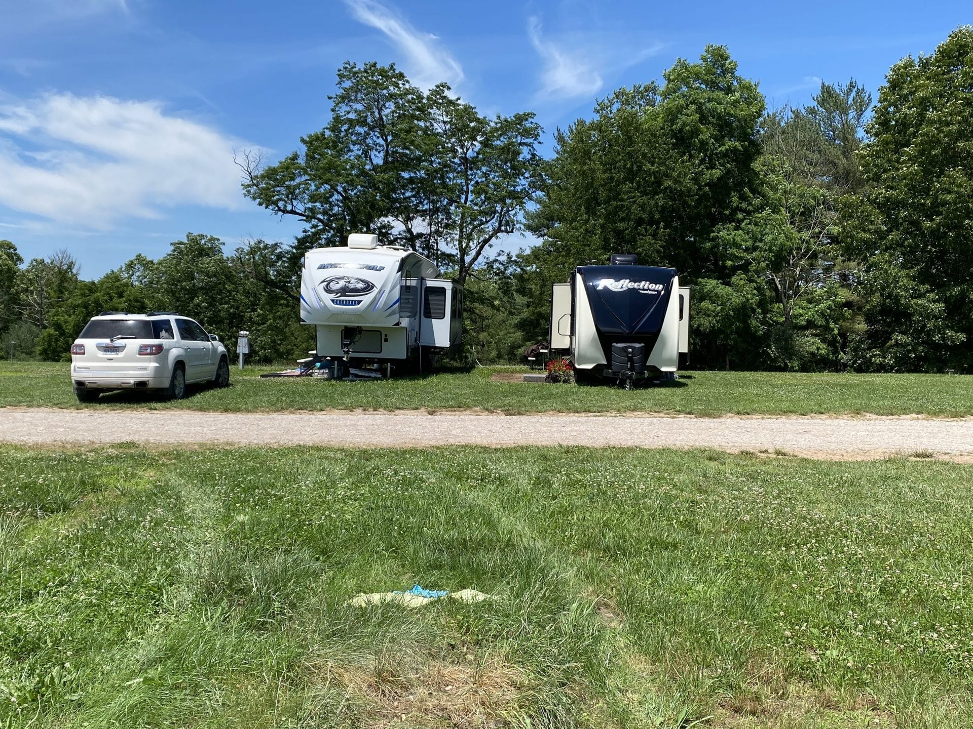 Two campers and a white SUV parked on a gravel drive near a grassy field under a sunny blue sky.