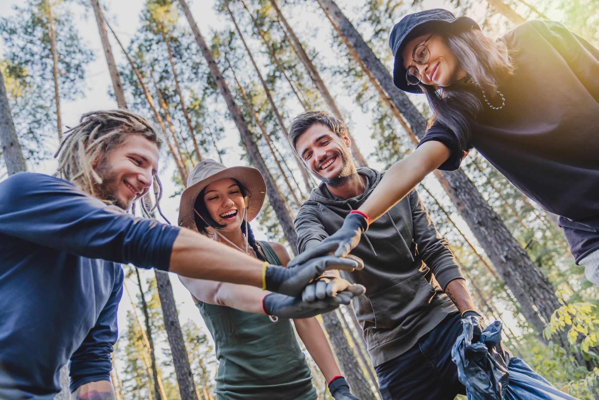 Four people wearing work gloves stack their hands in a forest, smiling while participating in an outdoor volunteer event.