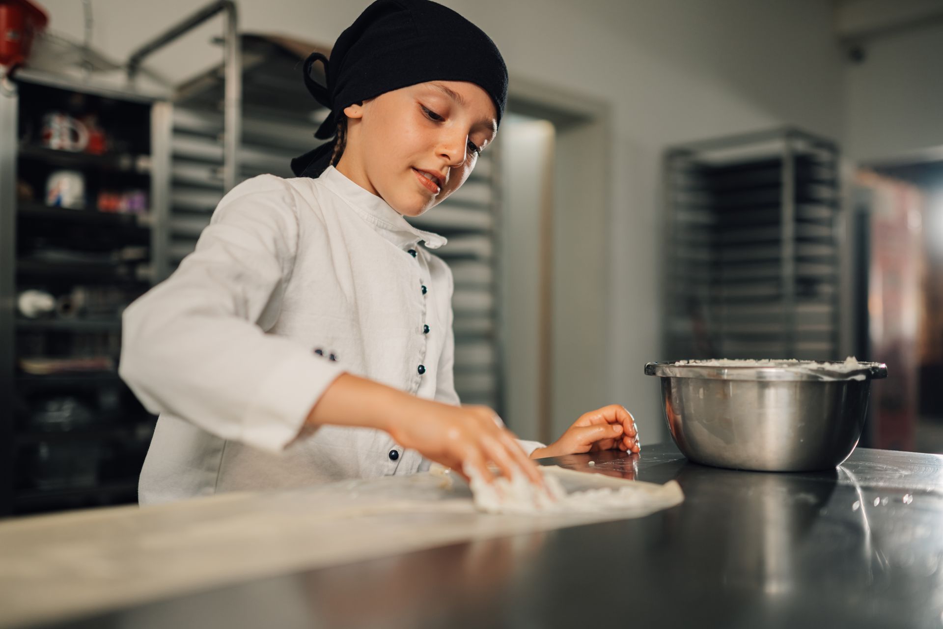 A baker in a white uniform and black cap sprinkles flour on a workspace, with a metal bowl nearby in a kitchen.