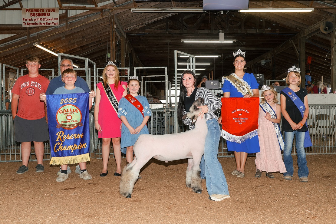 Two participants stand in an indoor livestock ring holding sheep while shaking hands with a judge in a fair setting.