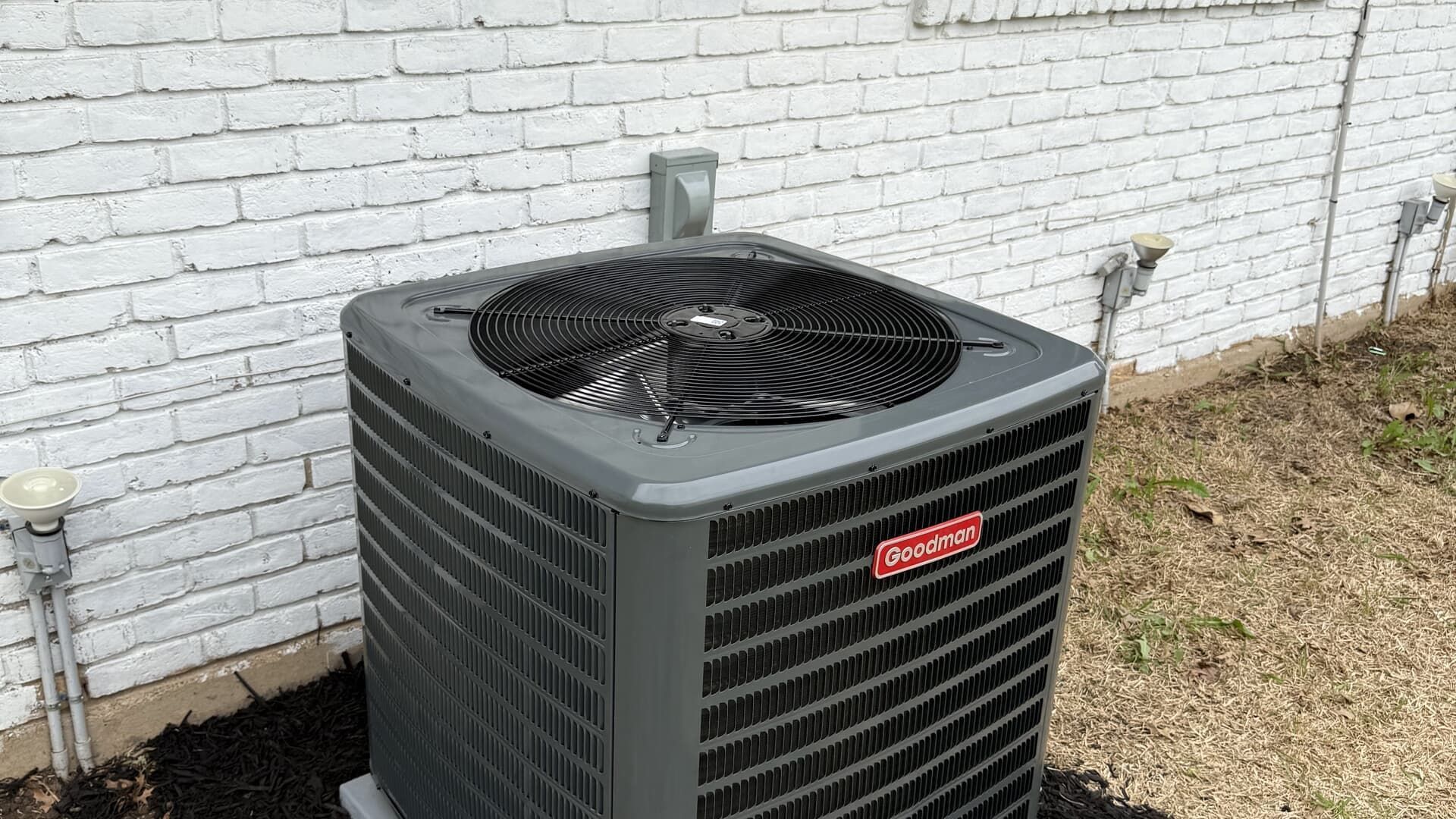 A grey Goodman central air conditioning unit sits outside on gravel against a white brick wall.