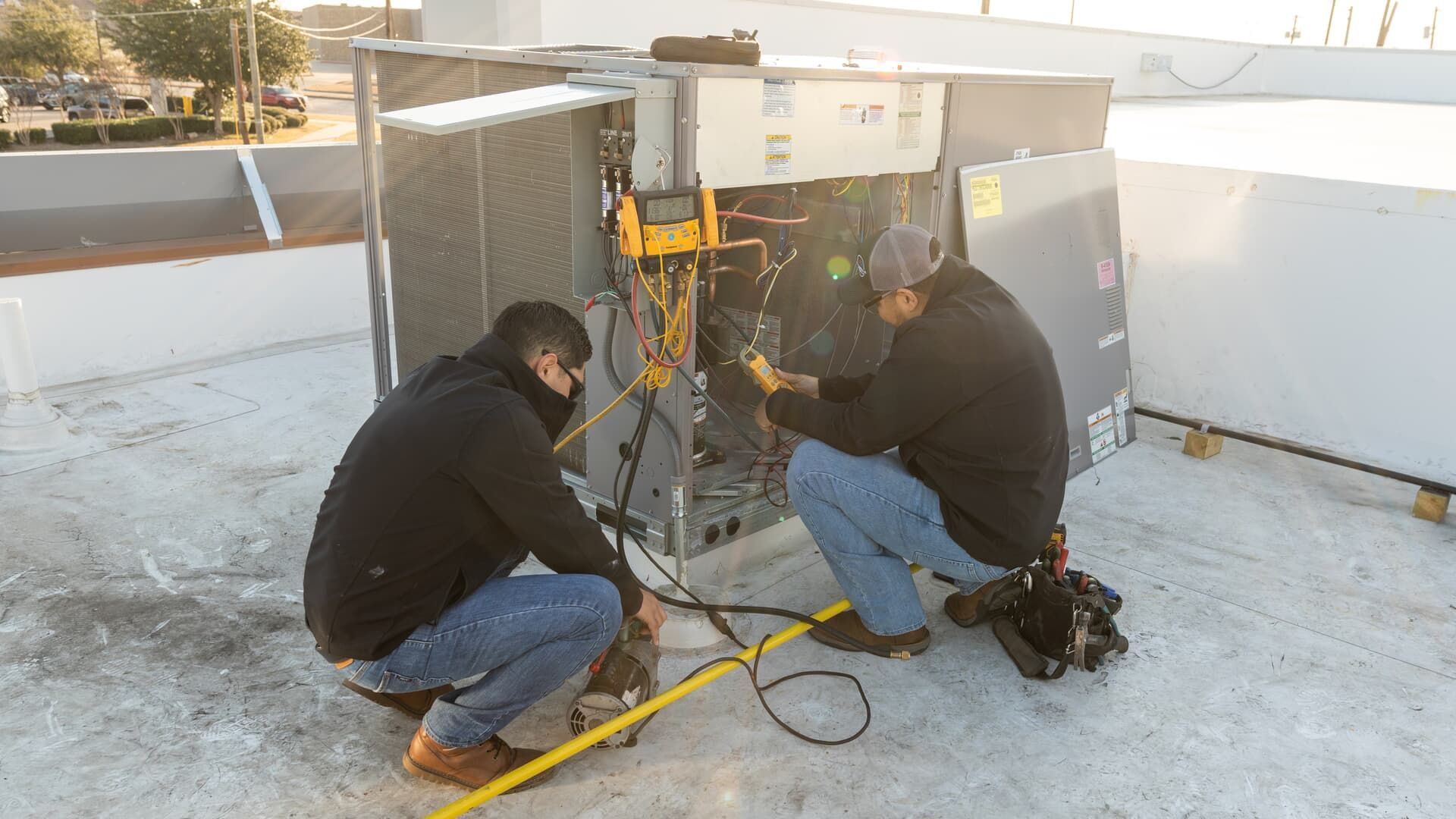 Two technicians in jackets and jeans service an HVAC unit on a flat rooftop, using diagnostic tools and wires.