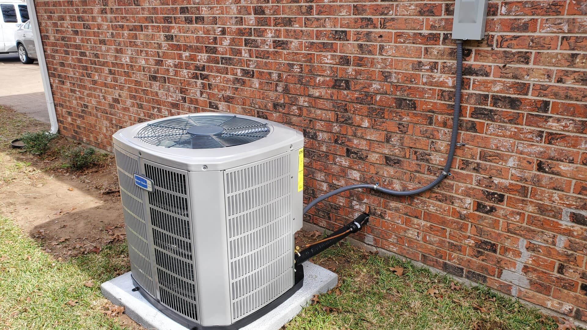 A modern, light gray HVAC outdoor unit sits on a concrete pad against a red brick wall with visible wiring and piping.