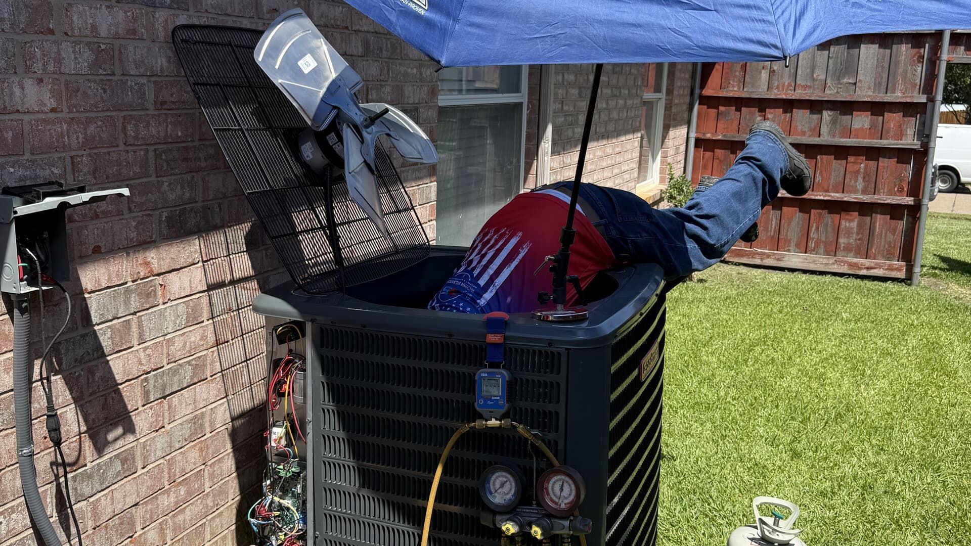 A technician works on an outdoor air conditioning unit while shielded by a blue umbrella.