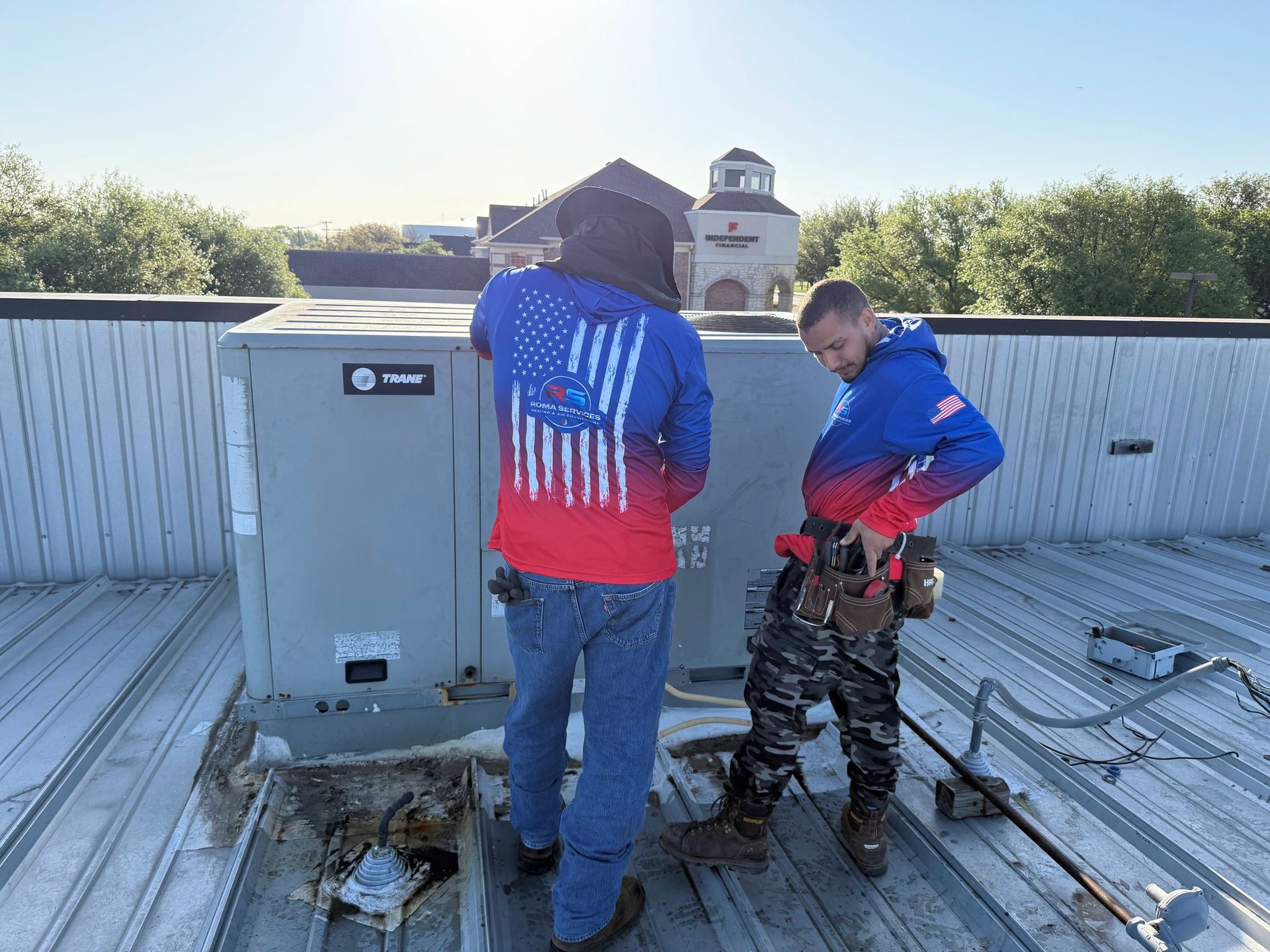 Two technicians wearing matching American flag hoodies service an HVAC unit on a commercial rooftop.