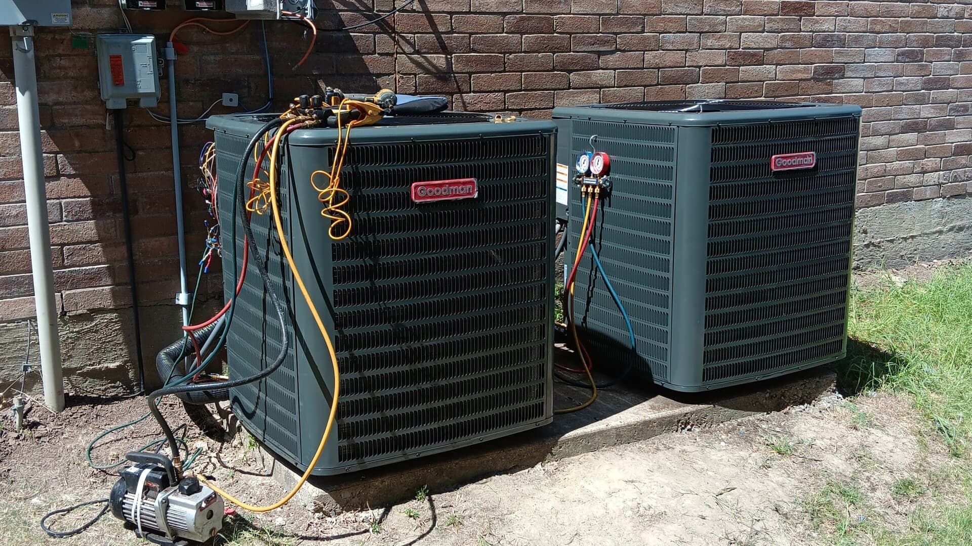 Two dark-gray HVAC units sit on a concrete pad against a brick wall, with a vacuum pump connected to one.