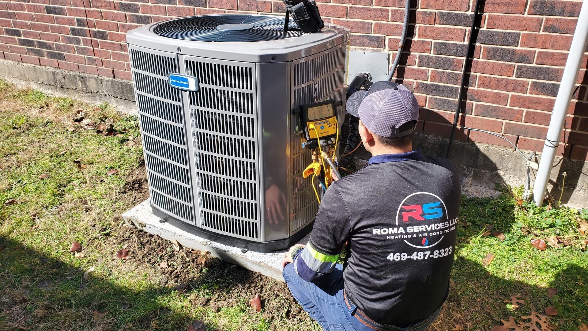 A technician wearing a branded work shirt services an outdoor air conditioning unit next to a brick wall.