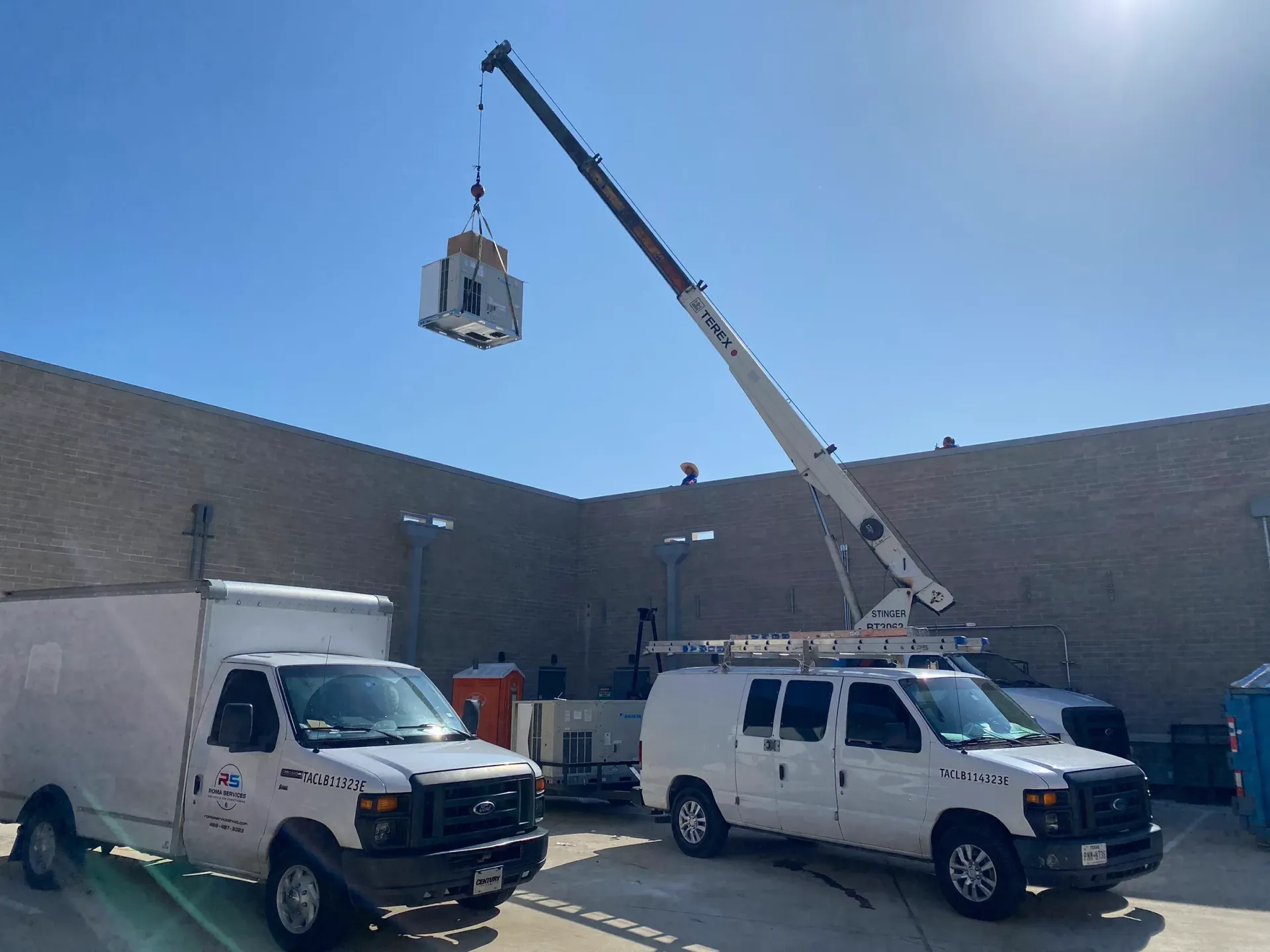 A crane truck lifts a piece of HVAC equipment over a building, with two white work vans parked below under a blue sky.