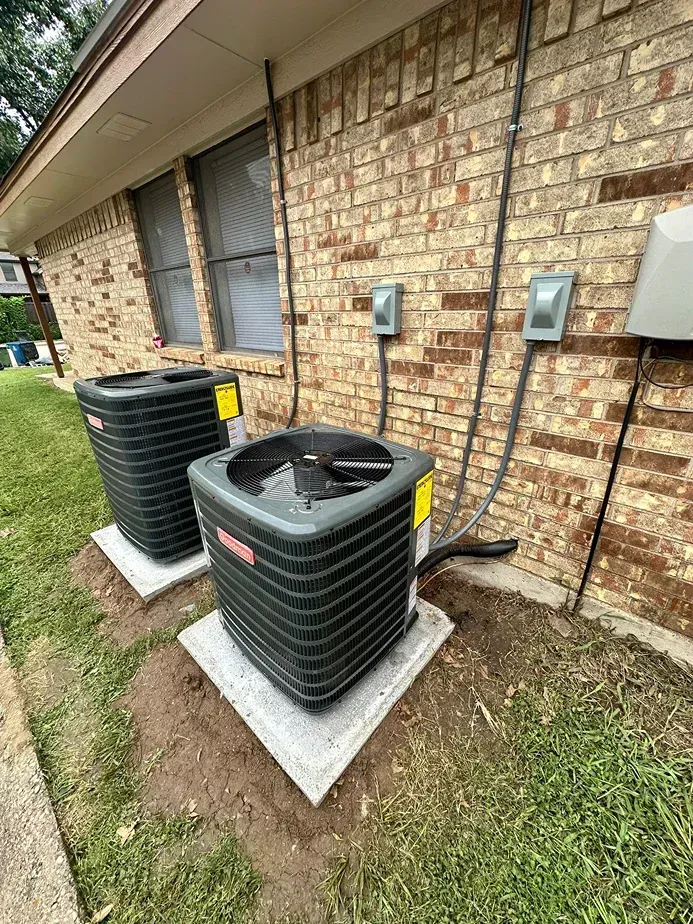 Two gray air conditioning units sitting on concrete pads against a brick exterior wall of a building.