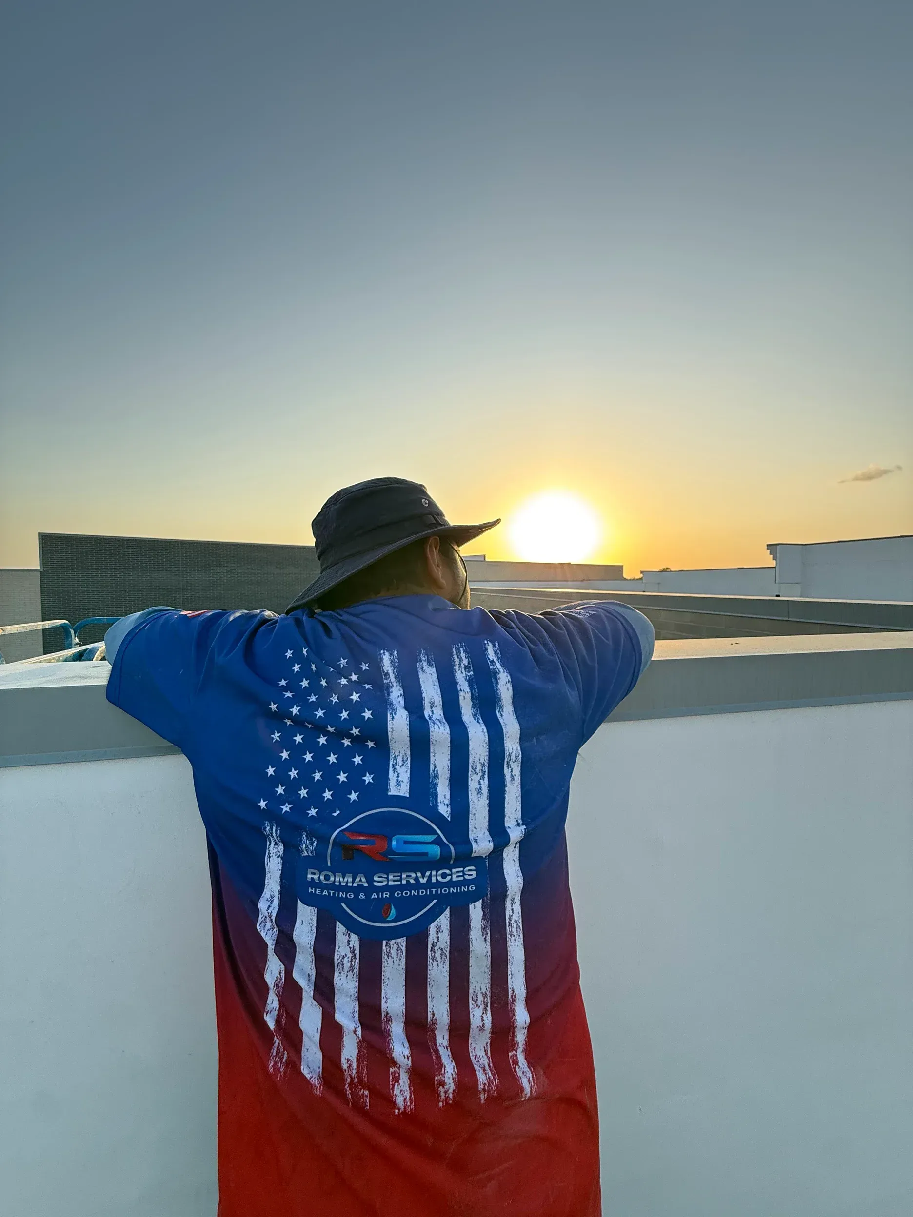 Person wearing a blue and red American flag shirt and a bucket hat, standing on a rooftop facing a golden sunrise.