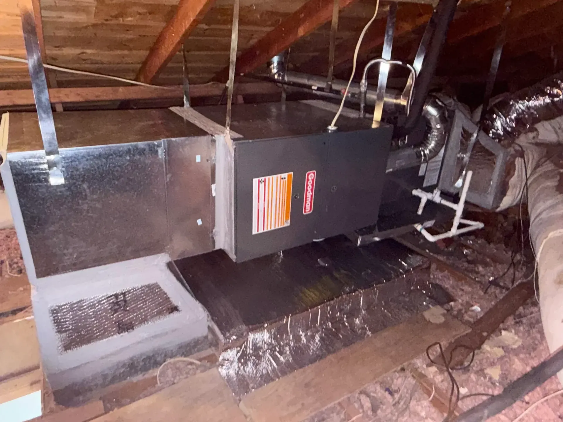 An HVAC air handler unit suspended by metal straps in a wooden attic space, connected to insulated silver ductwork.