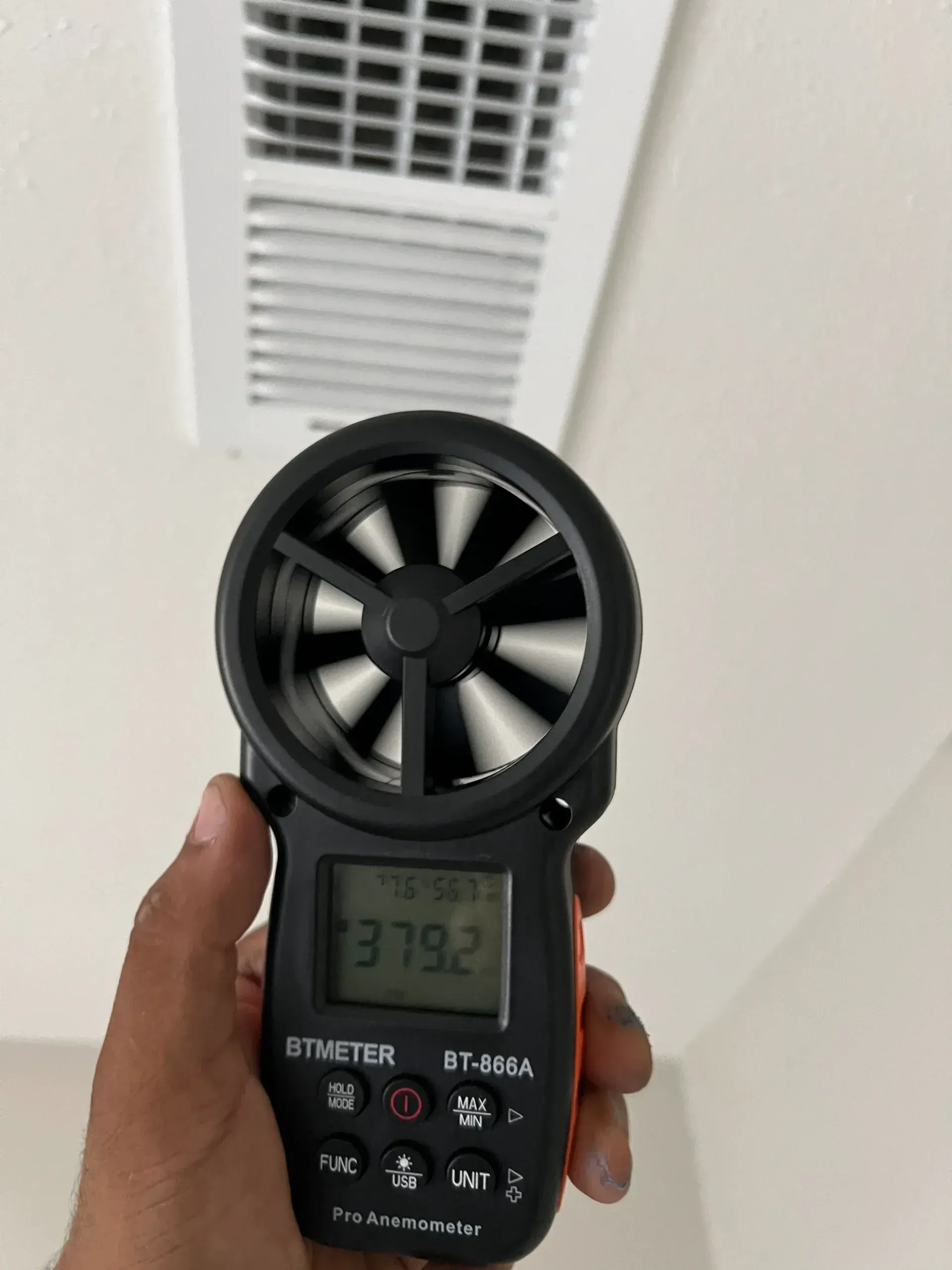 A hand holds a digital anemometer measuring air speed at 379.2 FPM in front of a ceiling HVAC vent.