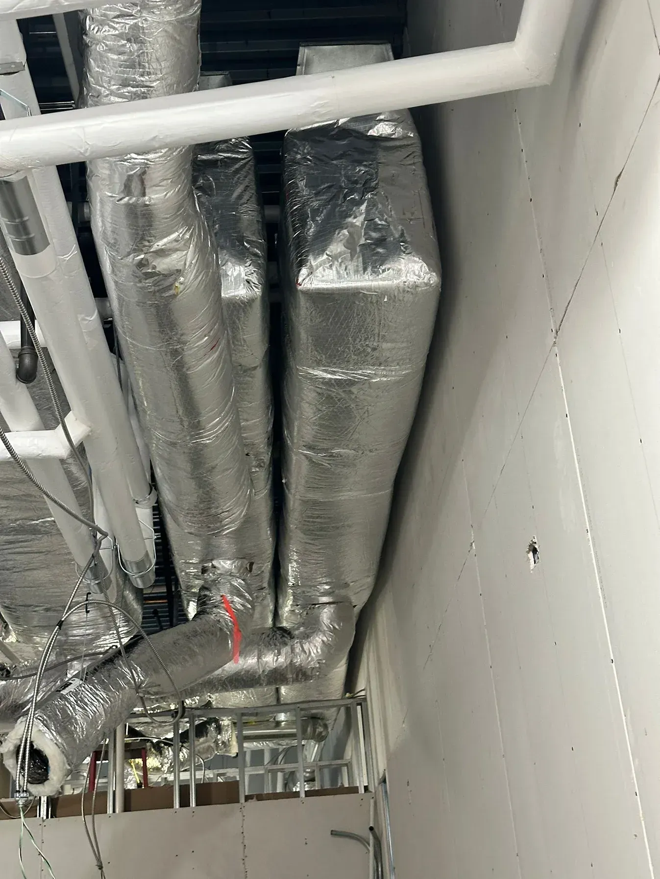 Metallic, foil-wrapped rectangular HVAC ducts and pipes running along a ceiling near a wall.