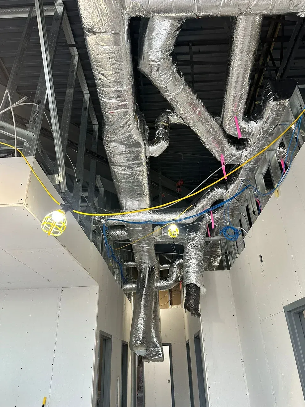 An unfinished hallway showing exposed silver ductwork, metal ceiling framing, and yellow work lights in a construction site.