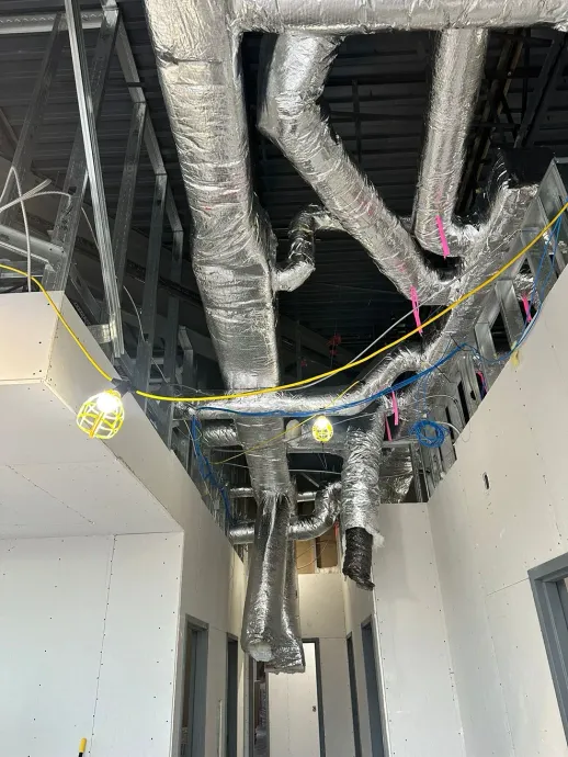 A construction hallway with unfinished white walls, exposed ceiling metal studs, silver air ducts, and hanging lights.
