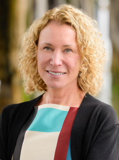 Portrait of a smiling person in a black jacket and colorful blouse, outdoors with a blurred background